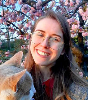 Photo of me, SachiiA Draws, a white girl with long dark blond hair, smiling brightly while holding up a cat. Blossoms are in the background.